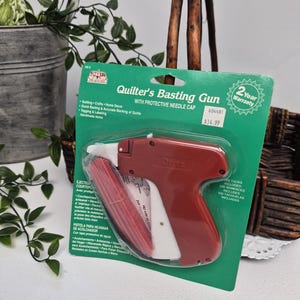 May include: A Dritz brand Quilter's Basting Gun with a protective needle cap, packaged on a green cardboard backing. The tool is red and white, designed for quilting, crafts, and home decor, and includes 500 tacks for tagging and labelling.