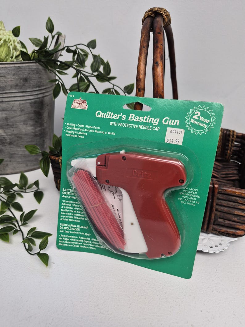 May include: A Dritz brand Quilter's Basting Gun with a protective needle cap, packaged on a green cardboard backing. The tool is red and white, designed for quilting, crafts, and home decor, and includes 500 tacks for tagging and labelling.