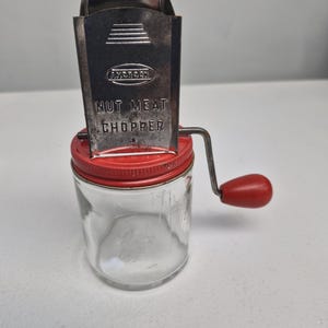 May include: A vintage nut and meat chopper with a clear glass jar and a red lid. The metal chopper has a crank handle with a red knob. The text "NUT MEAT CHOPPER" is printed on the front.