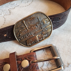 以下が含まれることがあります： 都市景観のデザインが施された、建物、クレーン、トラックなどを含む詳細な真鍮バックルが付いた茶色の革ベルト。ベルトには装飾的な花柄とシルバーカラーのバックルが付いています。
