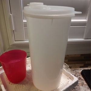 May include: A white plastic jug with a lid and a red plastic cup on a white lace doily.