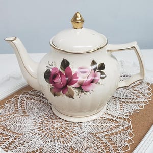 May include: Cream ceramic teapot with gold lid and handle details. The teapot is adorned with pink and red roses and green leaves. It rests on a white lace doily, a classic tea set item.
