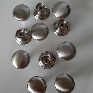 K&ouml;nnte beinhalten: Zehn silberne Schrankkn&ouml;pfe mit geb&uuml;rsteter Oberfl&auml;che. Einige Kn&ouml;pfe haben ein erhabenes kreisf&ouml;rmiges Design, w&auml;hrend andere flach sind.