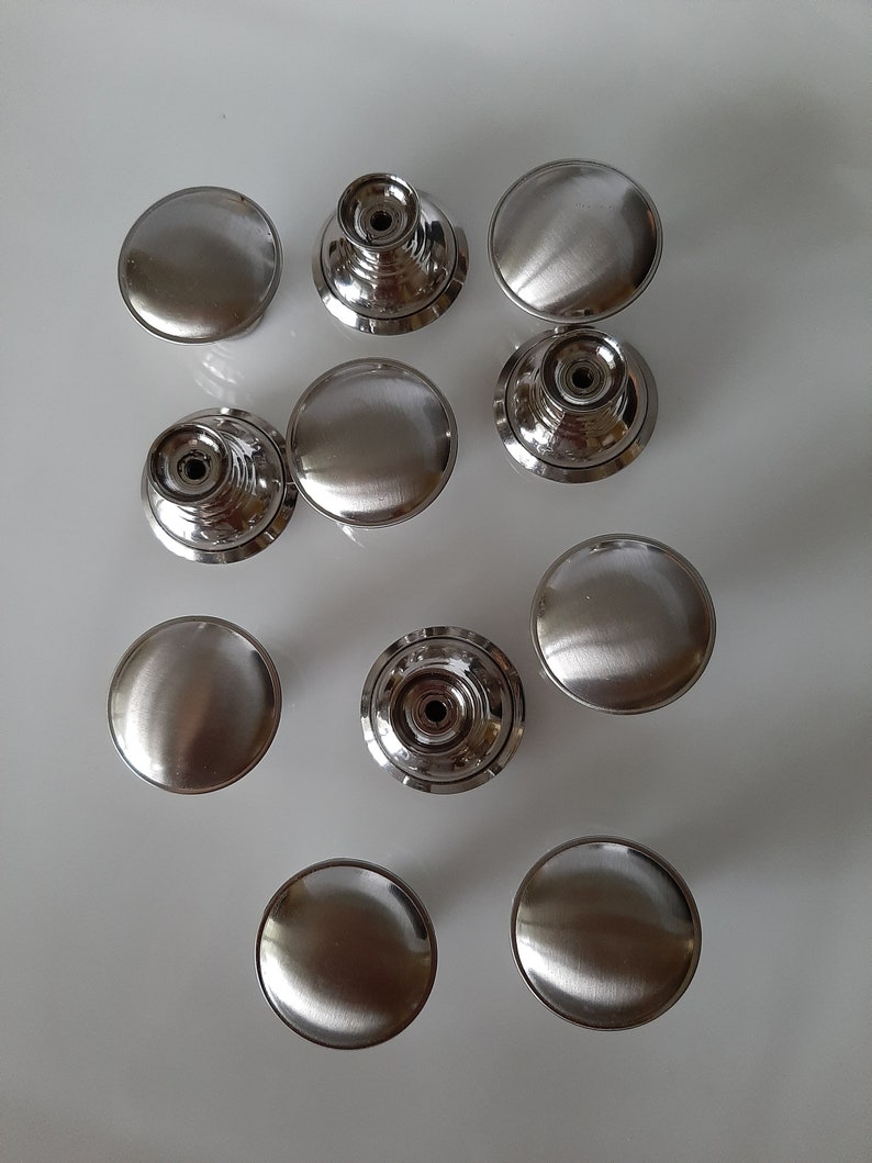 K&ouml;nnte beinhalten: Zehn silberne Schrankkn&ouml;pfe mit geb&uuml;rsteter Oberfl&auml;che. Einige Kn&ouml;pfe haben ein erhabenes kreisf&ouml;rmiges Design, w&auml;hrend andere flach sind.