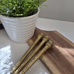 May include: Three cylindrical brass spindles with a ribbed, textured surface and a rounded, tiered top section. Each spindle features a consistent metallic gold finish and a tapered end, arranged diagonally in a parallel formation.