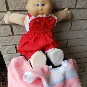 May include: A Cabbage Patch Kid doll wearing a red overall dress with white lace trim and white shoes. The doll is sitting on a pink and white striped sweater and a pink fuzzy sweater.