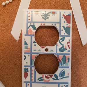 May include: A white ceramic outlet cover with a colorful tile pattern featuring houses, flowers, and geometric shapes. The tile pattern is in shades of blue, green, pink, and red.