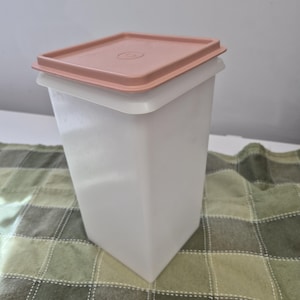 May include: A tall, rectangular storage container made of translucent white plastic with a flat, dusty rose-coloured lid. The lid features a centred circular indentation with a small embossed logo, and the container has a tapered base.