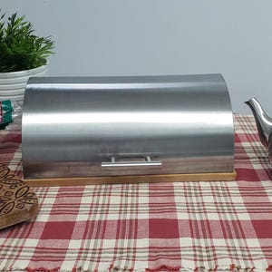 Peut inclure: Une boîte à pain en acier inoxydable avec une base en bois est posée sur une nappe à carreaux rouges et blancs. Une théière argentée avec un bouton noir et une petite plante en pot sont également sur la table. Une miche de pain est sur une planche à découper en bois.
