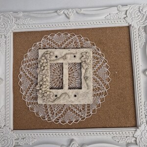 May include: A white ornate picture frame containing a brown cork board, a circular white lace doily, and a rectangular light beige light switch cover plate featuring a raised relief design of grape clusters and vine leaves on the surface.