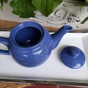 May include: A blue ceramic teapot with a matching lid. The teapot has a round body and a curved spout. The lid has a small knob on top.
