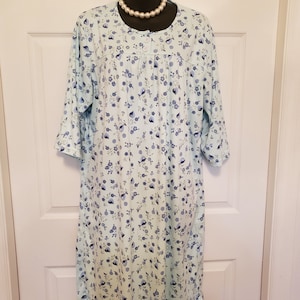 May include: A light blue nightgown with a floral pattern of blue and purple flowers. The nightgown has a round neckline, three-quarter sleeves, and a button-up front. A pearl necklace is around the mannequin's neck.
