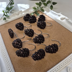 May include: A set of twelve dark brown, textured pinecone-shaped shower curtain hooks. Each individual hook features a detailed, realistic pinecone topper attached to a smooth, silver-toned metal curved hook designed for hanging curtains.