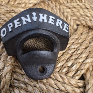 May include: A black cast iron wall-mounted bottle opener with a textured finish. The top curved section features the white embossed text "OPEN HERE" with a downward-pointing arrow between the words. It includes a circular hole for mounting.