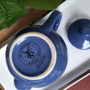 May include: A blue ceramic teapot with a lid and a windmill design. The teapot is labeled "Old Amsterdam 1701 Porcelain Works Lead Free Microwave Safe Dishwasher Safe".