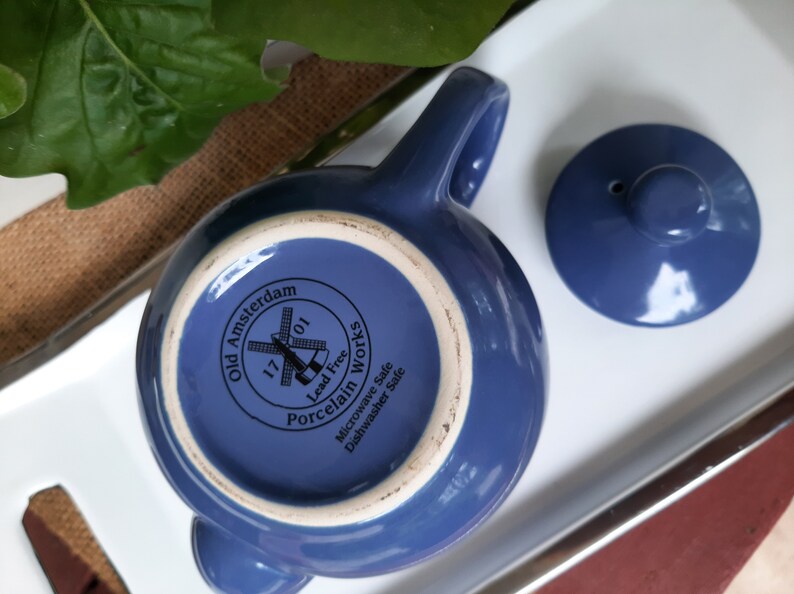 May include: A blue ceramic teapot with a lid and a windmill design. The teapot is labeled "Old Amsterdam 1701 Porcelain Works Lead Free Microwave Safe Dishwasher Safe".