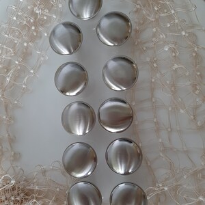 K&ouml;nnte beinhalten: Zehn silberfarbene runde Schrankkn&ouml;pfe mit glatter, gl&auml;nzender Oberfl&auml;che. Die Kn&ouml;pfe sind in zwei Reihen zu f&uuml;nf angeordnet, auf einer wei&szlig;en Oberfl&auml;che mit einem beigefarbenen Netzstoff als Hintergrund.