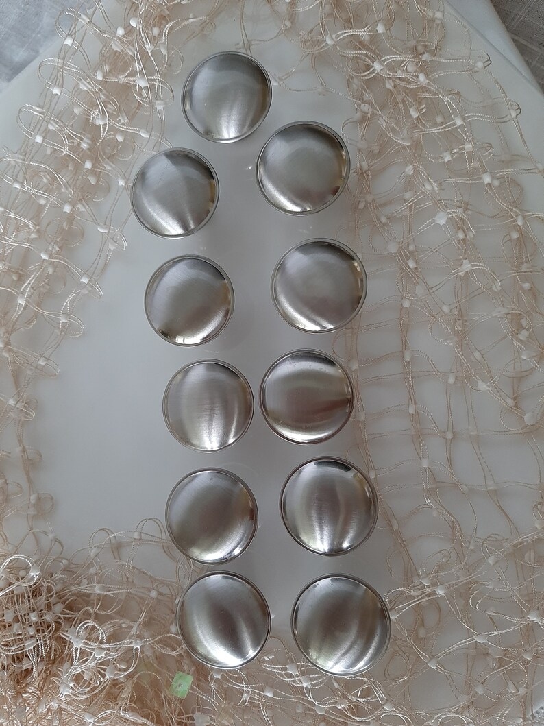 K&ouml;nnte beinhalten: Zehn silberfarbene runde Schrankkn&ouml;pfe mit glatter, gl&auml;nzender Oberfl&auml;che. Die Kn&ouml;pfe sind in zwei Reihen zu f&uuml;nf angeordnet, auf einer wei&szlig;en Oberfl&auml;che mit einem beigefarbenen Netzstoff als Hintergrund.