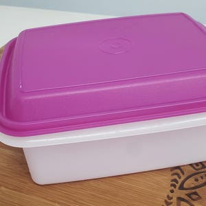 Könnte beinhalten: Ein rechteckiger Behälter aus weißem Kunststoff mit einem violetten Deckel. Der Deckel hat einen kleinen Kreis, der oben eingeprägt ist.