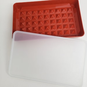 以下が含まれることがあります： 赤いプラスチック製の容器と透明な蓋。容器は小さなピラミッドのグリッドでテクスチャ加工された底面を持っています。蓋は半透明で、滑らかな表面をしています。