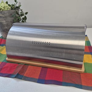 May include: A stainless steel bread box with a roll-top lid and a row of small ventilation holes on the front. The container features a rectangular light-colored wooden base and a clear plastic handle along the top edge of the curved metal cover.