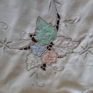 May include: White linen tablecloth with delicate embroidered floral design featuring a green leaf, a light blue flower, and a pink flower.