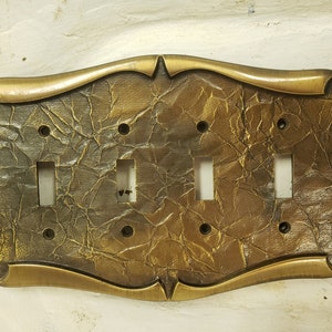 May include: A decorative light switch plate with a textured, crumpled appearance. The plate has a bronze finish and features four switch openings and a decorative border. The plate is designed to cover four light switches.