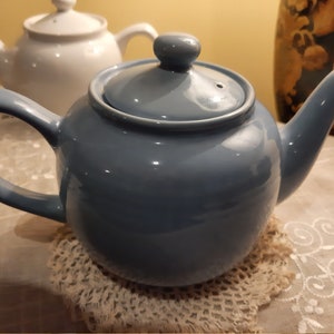 May include: A light blue ceramic teapot with a lid and a handle. The teapot is sitting on a white crocheted doily.