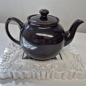 May include: A dark brown ceramic teapot with a matching lid, featuring a rounded body, a curved handle, and a tapered spout. The teapot has a glossy finish and is positioned on a white, ornate, rectangular frame with intricate relief detailing.