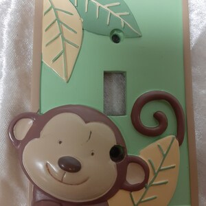 May include: A light green and brown plastic light switch cover with a monkey face and leaves. The monkey has a brown face with a smile and brown eyes. The leaves are green and yellow.
