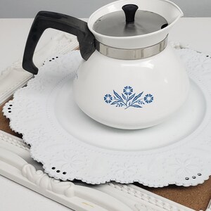 Op de afbeelding: Witte theepot van keramiek met een blauw bloemenmotief en een zwarte handgreep. De theepot staat op een wit kantonderzetter.