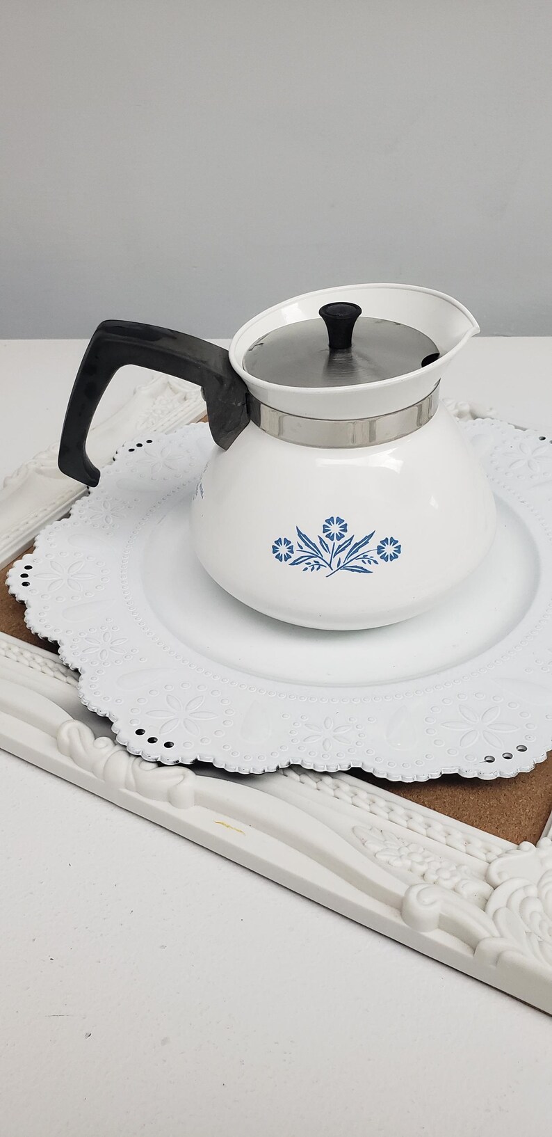 Op de afbeelding: Witte theepot van keramiek met een blauw bloemenmotief en een zwarte handgreep. De theepot staat op een wit kantonderzetter.