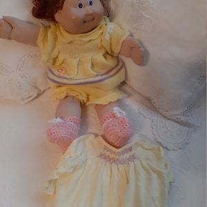 May include: A Cabbage Patch Kid doll with red curly hair, blue eyes, and a yellow knitted dress. The doll is wearing pink knitted booties and is sitting on a white bedspread.