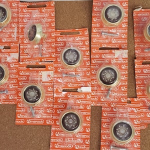 Pode incluir: Uma coleção de 12 botões de armário dourados com desenhos florais pretos. Os botões são embalados em sacos de plástico individuais com a marca "Amerock" impressa na embalagem.