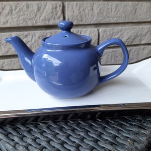 May include: A blue ceramic teapot with a rounded body and a lid with a small knob on top. The teapot is sitting on a white tray with a silver trim.