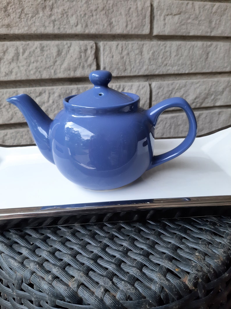 May include: A blue ceramic teapot with a rounded body and a lid with a small knob on top. The teapot is sitting on a white tray with a silver trim.