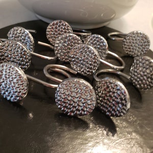 以下が含まれることがあります： テクスチャ加工が施された丸いデザインのシルバーのシャワーカーテンリング12個セット。リングは黒い表面に並べてあります。