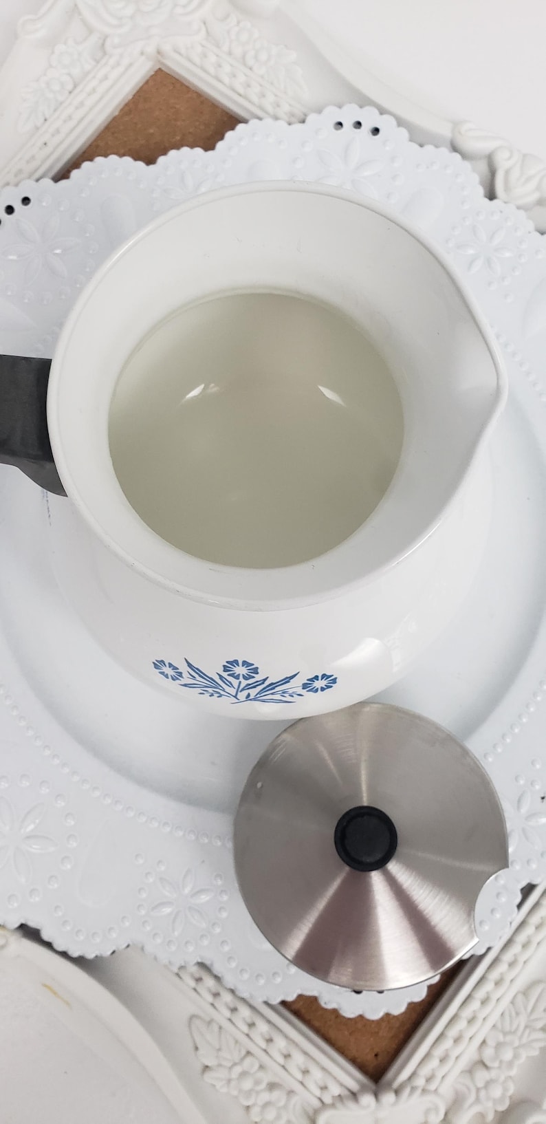 Op de afbeelding: Witte theepot van keramiek met een blauw bloemenmotief en een deksel van roestvrij staal. De theepot staat op een wit kantje.