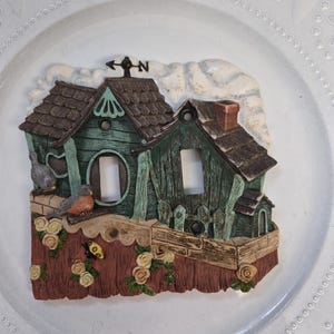 May include: A decorative light switch cover depicting a rustic village scene. The cover features two houses with brown shingled roofs, green walls, and a small chimney. A bird, a butterfly, and flowers add detail.