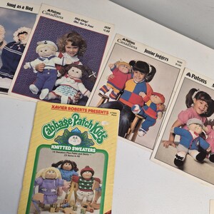 Peut inclure: Une collection de cinq livrets de modèles de tricot vintage Cabbage Patch Kids par Patons, incluant les titres Snug as a Bug, Ship Ahoy, Junior Joggers, All Dressed Up et Knitted Sweaters, montrant diverses poupées dans des tenues tricotées.