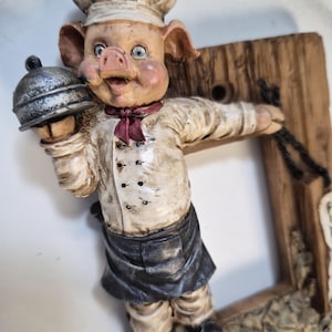 May include: A whimsical figurine of a pig dressed as a chef, holding a covered dish. The pig wears a white chef's hat, jacket, and a black apron. The figurine is attached to a wooden frame, adding a rustic touch to the design.