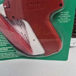 May include: A red and white Dritz tagging gun tool for attaching labels to fabric, packaged in a green blister pack. The packaging includes the text 500 TACKS INCLUDED, 500 SEMENCES INCLUSES, and 500 TACHUELAS INCLUIDAS, along with a red plastic needle cover.