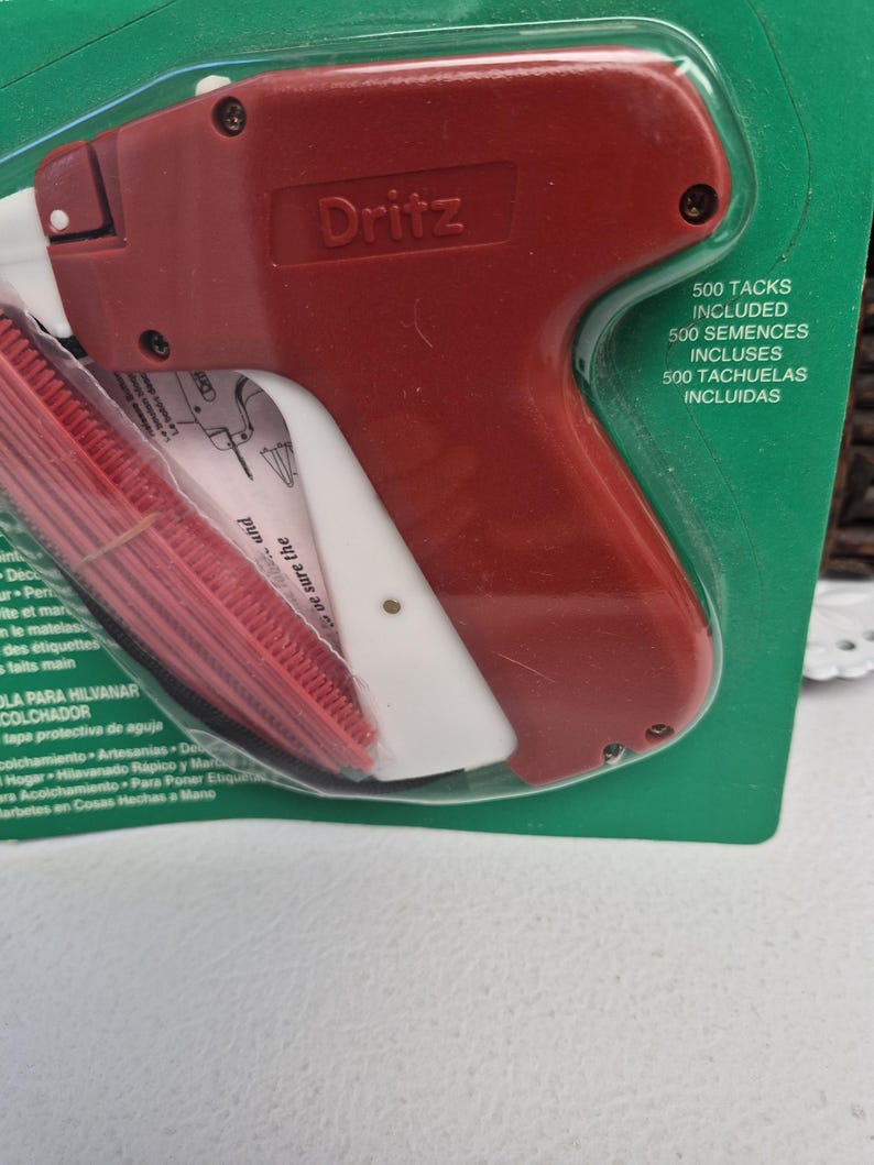 May include: A red and white Dritz tagging gun tool for attaching labels to fabric, packaged in a green blister pack. The packaging includes the text 500 TACKS INCLUDED, 500 SEMENCES INCLUSES, and 500 TACHUELAS INCLUIDAS, along with a red plastic needle cover.