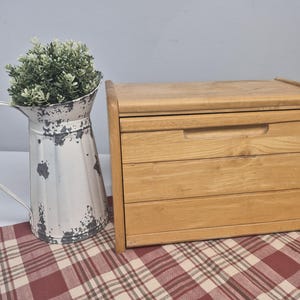 Peut inclure: Un coffre à pain en bois clair avec un dessus incurvé et une poignée, à côté d'une cruche en métal blanc vieilli remplie de verdure. Le coffre à pain a trois tiroirs visibles. Sur un tissu à carreaux rouges et blancs.
