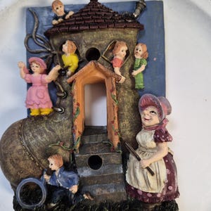 May include: A whimsical, decorative boot-shaped figurine with a house built into it. The boot is brown and houses several small figures of children. A woman in a bonnet stands nearby. The background is blue.