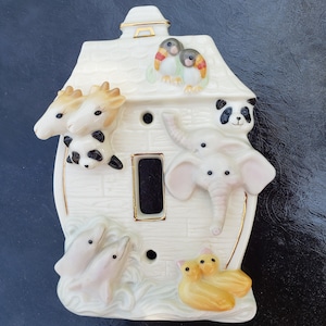 May include: A white ceramic light switch cover with a house design featuring various animals, including a giraffe, panda, elephant, dolphins, and birds. The house has a gold trim and a brown roof.
