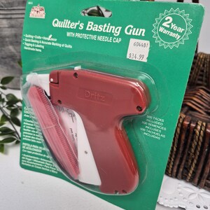 May include: A Dritz brand Quilter's Basting Gun with a protective needle cap, packaged on a green cardboard backing. The tool is dark red with a white trigger and handle. The packaging includes text, a 2-year warranty seal, and 500 tacks included.