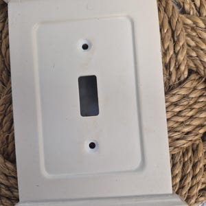 Puede incluir: Cubierta de interruptor de luz rectangular de color blanquecino con un diseño decorativo enmarcado. La cubierta tiene una abertura para un interruptor y dos orificios para tornillos. Fabricada con un material sólido y con un acabado ligeramente envejecido.