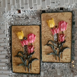 May include: Two decorative wall plaques with a 3D design of tulips. The plaques are made of a textured material and feature a light brown background with green leaves and yellow and pink tulips.