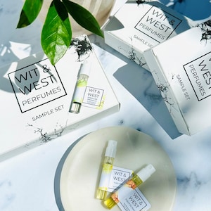 May include: A flat lay of perfume sample sets. White boxes with black marble-like accents and the text "WIT & WEST PERFUMES SAMPLE SET" are visible. Small glass vials of perfume and labels with fragrance names are also present. Green leaves and a white plate add to the aesthetic.